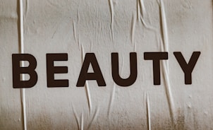 Large, bold letters spelling the word 'BEAUTY' are displayed on a textured surface. The background appears to have a subtle beige tint with visible creases or folds.