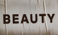 Large, bold letters spelling the word 'BEAUTY' are displayed on a textured surface. The background appears to have a subtle beige tint with visible creases or folds.