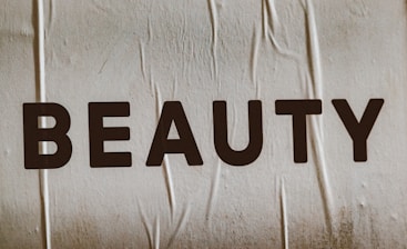 Large, bold letters spelling the word 'BEAUTY' are displayed on a textured surface. The background appears to have a subtle beige tint with visible creases or folds.