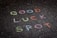 Colorful chalk words are written on a dark, hexagonal-patterned pavement. The words read 'GOOD LUCK SPOT.' Each word is a different color, providing a contrast to the dark surface.