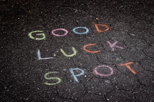 Colorful chalk words are written on a dark, hexagonal-patterned pavement. The words read 'GOOD LUCK SPOT.' Each word is a different color, providing a contrast to the dark surface.