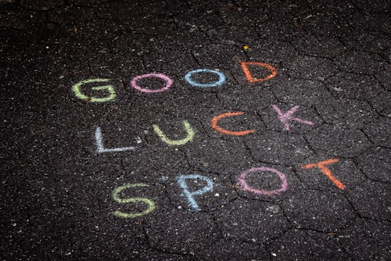 Colorful chalk words are written on a dark, hexagonal-patterned pavement. The words read 'GOOD LUCK SPOT.' Each word is a different color, providing a contrast to the dark surface.