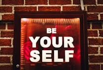 A framed sign with the words 'BE YOUR SELF' in bold white letters is set against a dark red and orange background. The sign is mounted on a brick wall which is visible around the edges.