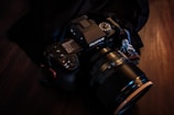 A black Fujifilm camera with a large lens is placed on a dark surface, surrounded by a black cloth. The camera displays settings on its screen and has various dials and buttons, indicating it is a professional or semi-professional model.