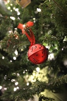 A red ornament with the word 'Joy' and decorative patterns hangs from a branch of a Christmas tree. The ornament is tied with a red ribbon, and surrounding the ornament are the green needles of the tree. The background is blurred with small, glowing lights, creating a festive atmosphere.