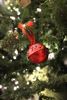 A red ornament with the word 'Joy' and decorative patterns hangs from a branch of a Christmas tree. The ornament is tied with a red ribbon, and surrounding the ornament are the green needles of the tree. The background is blurred with small, glowing lights, creating a festive atmosphere.