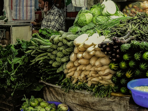 A market stall displaying an assortment of fresh vegetables including green leafy produce, white radishes, eggplants, luffa, and a cabbage in the background. A vendor stands at the side, possibly attending to customers. The vegetables are neatly arranged in piles with a blue container seen at the bottom right.