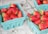 Several cartons of fresh strawberries are neatly arranged in teal-colored baskets, sitting on a textured surface. The strawberries are bright red with green stems, and they appear freshly picked.