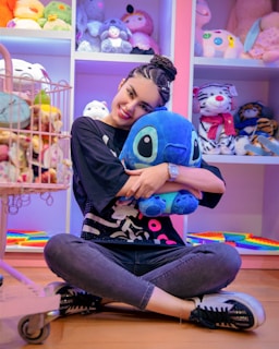 A person is sitting on the floor, smiling and embracing a large blue plush toy in a store filled with various colorful stuffed animals. Shelves and displays around are filled with diverse plush toys, creating a playful and whimsical atmosphere.