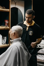 A barber carefully trims the hair of a seated customer with short, light-blonde hair. The barber is wearing a black polo shirt with a yellow collar, and seems focused on his work. In the background, shelves hold various grooming products.