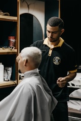 A barber carefully trims the hair of a seated customer with short, light-blonde hair. The barber is wearing a black polo shirt with a yellow collar, and seems focused on his work. In the background, shelves hold various grooming products.