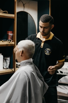 A barber carefully trims the hair of a seated customer with short, light-blonde hair. The barber is wearing a black polo shirt with a yellow collar, and seems focused on his work. In the background, shelves hold various grooming products.