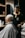 A barber carefully trims the hair of a seated customer with short, light-blonde hair. The barber is wearing a black polo shirt with a yellow collar, and seems focused on his work. In the background, shelves hold various grooming products.