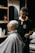 A barber carefully trims the hair of a seated customer with short, light-blonde hair. The barber is wearing a black polo shirt with a yellow collar, and seems focused on his work. In the background, shelves hold various grooming products.