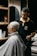 A barber carefully trims the hair of a seated customer with short, light-blonde hair. The barber is wearing a black polo shirt with a yellow collar, and seems focused on his work. In the background, shelves hold various grooming products.