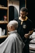 A barber carefully trims the hair of a seated customer with short, light-blonde hair. The barber is wearing a black polo shirt with a yellow collar, and seems focused on his work. In the background, shelves hold various grooming products.