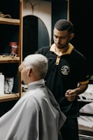 A barber carefully trims the hair of a seated customer with short, light-blonde hair. The barber is wearing a black polo shirt with a yellow collar, and seems focused on his work. In the background, shelves hold various grooming products.