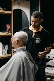 A barber carefully trims the hair of a seated customer with short, light-blonde hair. The barber is wearing a black polo shirt with a yellow collar, and seems focused on his work. In the background, shelves hold various grooming products.