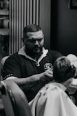 A barber with a beard and tattoos is giving a haircut to a customer. The barber is wearing a dark polo shirt with a logo on it. The scene takes place in a barbershop with a patterned wall in the background.