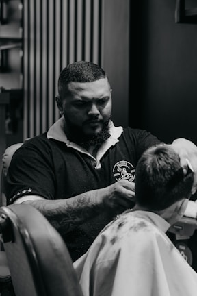A barber with a beard and tattoos is giving a haircut to a customer. The barber is wearing a dark polo shirt with a logo on it. The scene takes place in a barbershop with a patterned wall in the background.