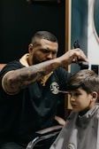 A barber is focused on cutting a young boy's hair, using clippers. The barber is wearing a black polo shirt with a yellow collar and a logo on the chest, and has tattoos on his arms. The boy, sitting in a barber chair covered with a cape, appears to be calm and somewhat serious.