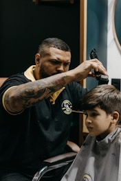 A barber is focused on cutting a young boy's hair, using clippers. The barber is wearing a black polo shirt with a yellow collar and a logo on the chest, and has tattoos on his arms. The boy, sitting in a barber chair covered with a cape, appears to be calm and somewhat serious.
