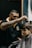 A barber is focused on cutting a young boy's hair, using clippers. The barber is wearing a black polo shirt with a yellow collar and a logo on the chest, and has tattoos on his arms. The boy, sitting in a barber chair covered with a cape, appears to be calm and somewhat serious.