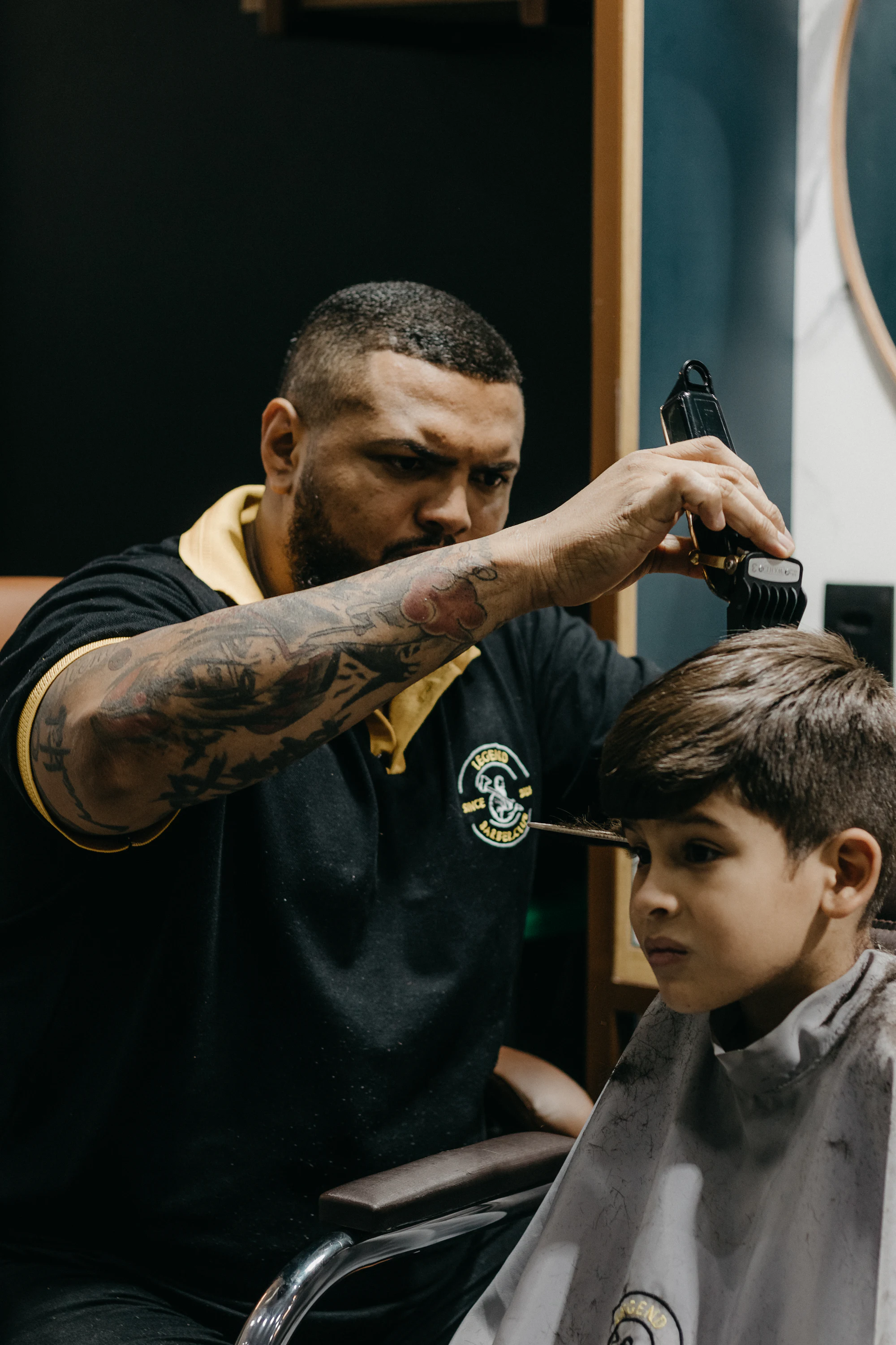 A stylist using clippers around the ear of a young boy sitting in a barber chair.