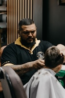 A barber is concentrated on cutting a client's hair in a modern barbershop. The barber has a tattooed arm and wears a black shirt with yellow accents. The client sits in a chair wearing a white cape with some cut hair visible on it. Wooden panels and other barbershop decor can be seen in the background.