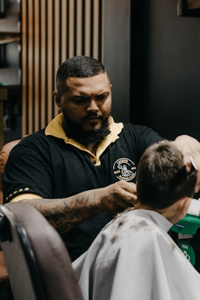 A barber is concentrated on cutting a client's hair in a modern barbershop. The barber has a tattooed arm and wears a black shirt with yellow accents. The client sits in a chair wearing a white cape with some cut hair visible on it. Wooden panels and other barbershop decor can be seen in the background.
