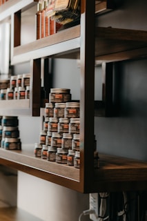Wooden shelves display rows of small jars and various bottles. The jars are stacked in a pyramid shape, each labeled, suggesting they contain products like creams or condiments. The rustic wooden design paired with the neatly arranged items creates an organized and artisanal appearance.