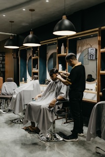 A modern barbershop interior with several styling stations. A person sits comfortably in a salon chair wearing a cape, while a barber is engaged in cutting their hair. The scene includes mirrors, shelves with products, and stylish lighting fixtures hanging from the ceiling.