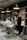 A modern barbershop interior with several styling stations. A person sits comfortably in a salon chair wearing a cape, while a barber is engaged in cutting their hair. The scene includes mirrors, shelves with products, and stylish lighting fixtures hanging from the ceiling.