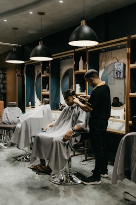A modern barbershop interior with several styling stations. A person sits comfortably in a salon chair wearing a cape, while a barber is engaged in cutting their hair. The scene includes mirrors, shelves with products, and stylish lighting fixtures hanging from the ceiling.