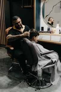 A barber is giving a haircut to a young boy who is sitting in a salon chair, draped with a white barber cape. The barber is focused on his work, with various tools and products scattered on the counter in front of a large round mirror. The background features dark walls with vertical wooden accents.