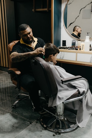 A barber is giving a haircut to a young boy who is sitting in a salon chair, draped with a white barber cape. The barber is focused on his work, with various tools and products scattered on the counter in front of a large round mirror. The background features dark walls with vertical wooden accents.