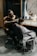 A barber is giving a haircut to a young boy who is sitting in a salon chair, draped with a white barber cape. The barber is focused on his work, with various tools and products scattered on the counter in front of a large round mirror. The background features dark walls with vertical wooden accents.