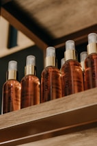Several spray bottles arranged neatly in a row on a wooden shelf. Each bottle is labeled with 'BELLA Blend Oil' and features a golden cap with a clear spray nozzle. The lighting creates a warm and inviting ambiance.
