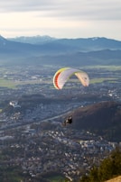 A paraglider soars over a picturesque landscape, with a vibrant parachute featuring stripes of multiple colors. Below, a vast cityscape unfolds, surrounded by lush green fields and distant mountains capped with snow.