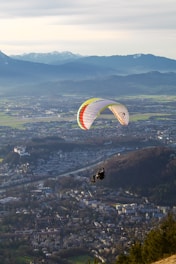 A paraglider soars over a picturesque landscape, with a vibrant parachute featuring stripes of multiple colors. Below, a vast cityscape unfolds, surrounded by lush green fields and distant mountains capped with snow.