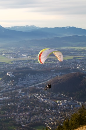 A paraglider soars over a picturesque landscape, with a vibrant parachute featuring stripes of multiple colors. Below, a vast cityscape unfolds, surrounded by lush green fields and distant mountains capped with snow.