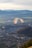 A paraglider soars over a picturesque landscape, with a vibrant parachute featuring stripes of multiple colors. Below, a vast cityscape unfolds, surrounded by lush green fields and distant mountains capped with snow.