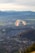 A paraglider soars over a picturesque landscape, with a vibrant parachute featuring stripes of multiple colors. Below, a vast cityscape unfolds, surrounded by lush green fields and distant mountains capped with snow.
