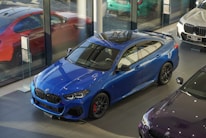 A sleek blue BMW car is displayed inside a modern showroom with large glass windows. Nearby, other cars in red, white, and a dark shade are visible, reflecting a contemporary and luxurious environment.