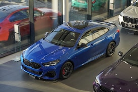 A sleek blue BMW car is displayed inside a modern showroom with large glass windows. Nearby, other cars in red, white, and a dark shade are visible, reflecting a contemporary and luxurious environment.