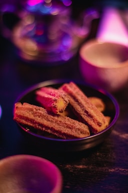 A bowl of churros dusted with sugar is placed on a dark surface, highlighted by warm lighting. In the background, there are blurred, colorful lights creating a cozy and inviting atmosphere.