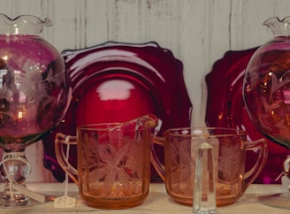 A collection of intricately designed glassware featuring floral patterns, set against a backdrop of deep red decorative plates. The items include two glass pitchers and an intricately detailed glass.