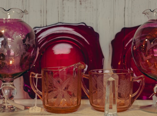 A collection of intricately designed glassware featuring floral patterns, set against a backdrop of deep red decorative plates. The items include two glass pitchers and an intricately detailed glass.