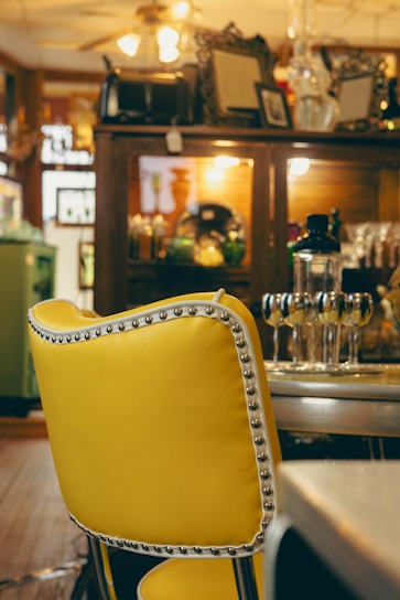 A brightly colored yellow chair with nailhead trim sits in the foreground. In the background, there is a wooden cabinet displaying various decorative items, including glassware and framed art. Overhead lighting casts a warm glow throughout the room.