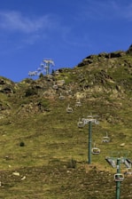 A series of chairlifts ascending a grassy, rocky hillside with a vibrant blue sky overhead. The chairlifts are unoccupied and spread in a line, supported by several tall metal towers.